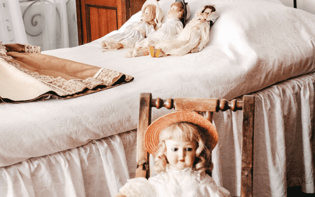 This is four white porcelain dolls on a bed in a bed in a room. Three dolls are on the bed which is adorned with white bed sheets and a cloth material folded at the center of the bed. The fourth doll is in a wooden chair facing towards the audience. This represent the eerie feeling of the characters in "Sharp Objects" and also, the use of dolls to accentuate horror.