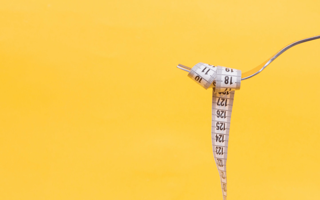 This is a fork with a measuring tape wrapped around the fork against a yellow background, representing the pressure for weight loss in "Shrill".