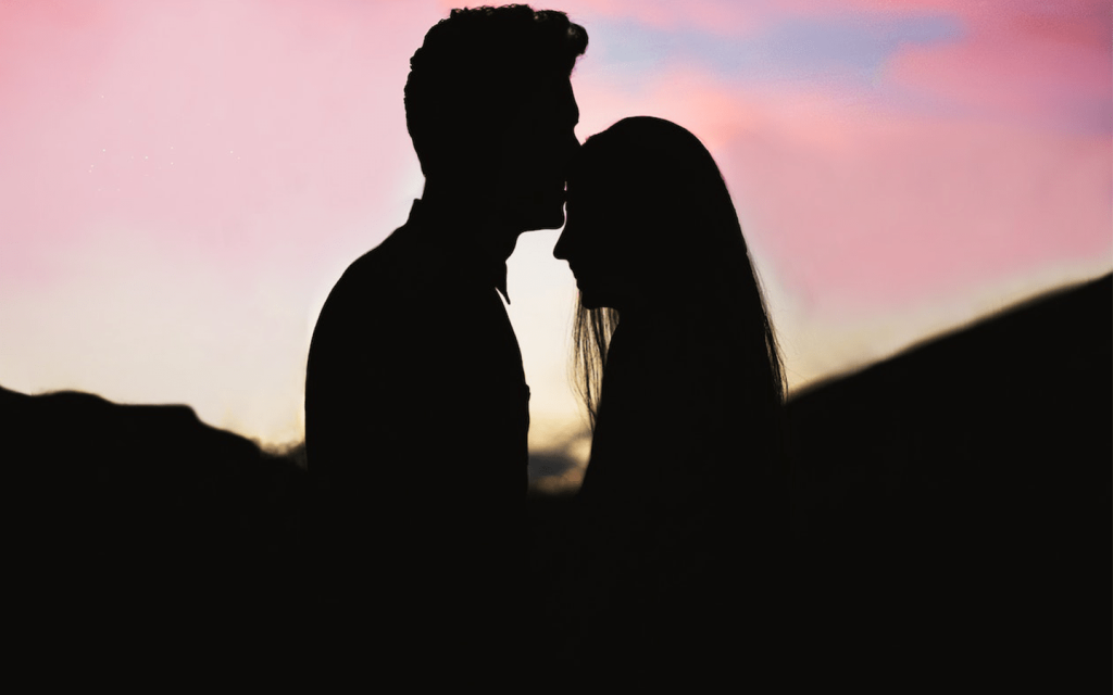 This is a darkened picture of a man kissing a woman's forehead againsst a pinkish sky. This represents Connell and Marianne in "Normal People".