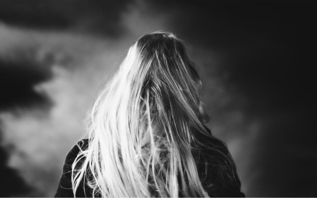 This is a black and white image of the back of a blonde woman with messy long hair looking towards an indistinguishable path which looks a lot like the back of Cassie in "Promising Young Woman".