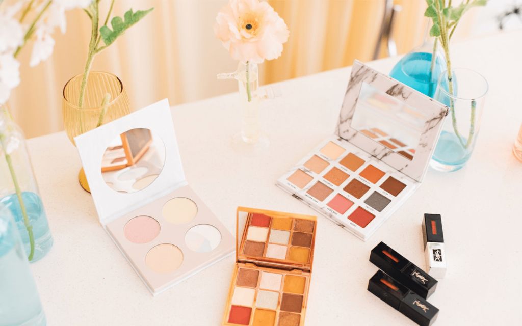 The is white tabke with eye shadow, lipstick, and vases with flowers with a light orange curtain at the background, to represent the makeup in "True Beauty".